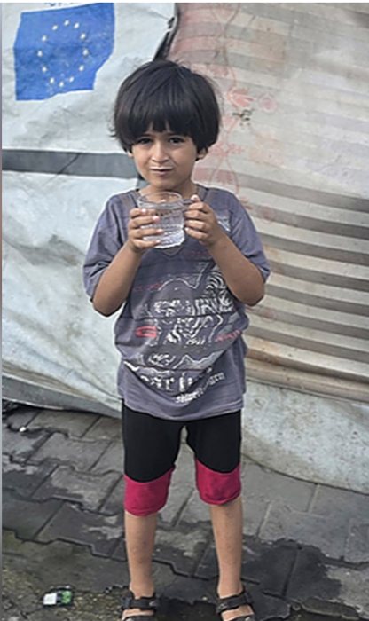 Hamza, aged 5, holding a cup of water outside the family's tent